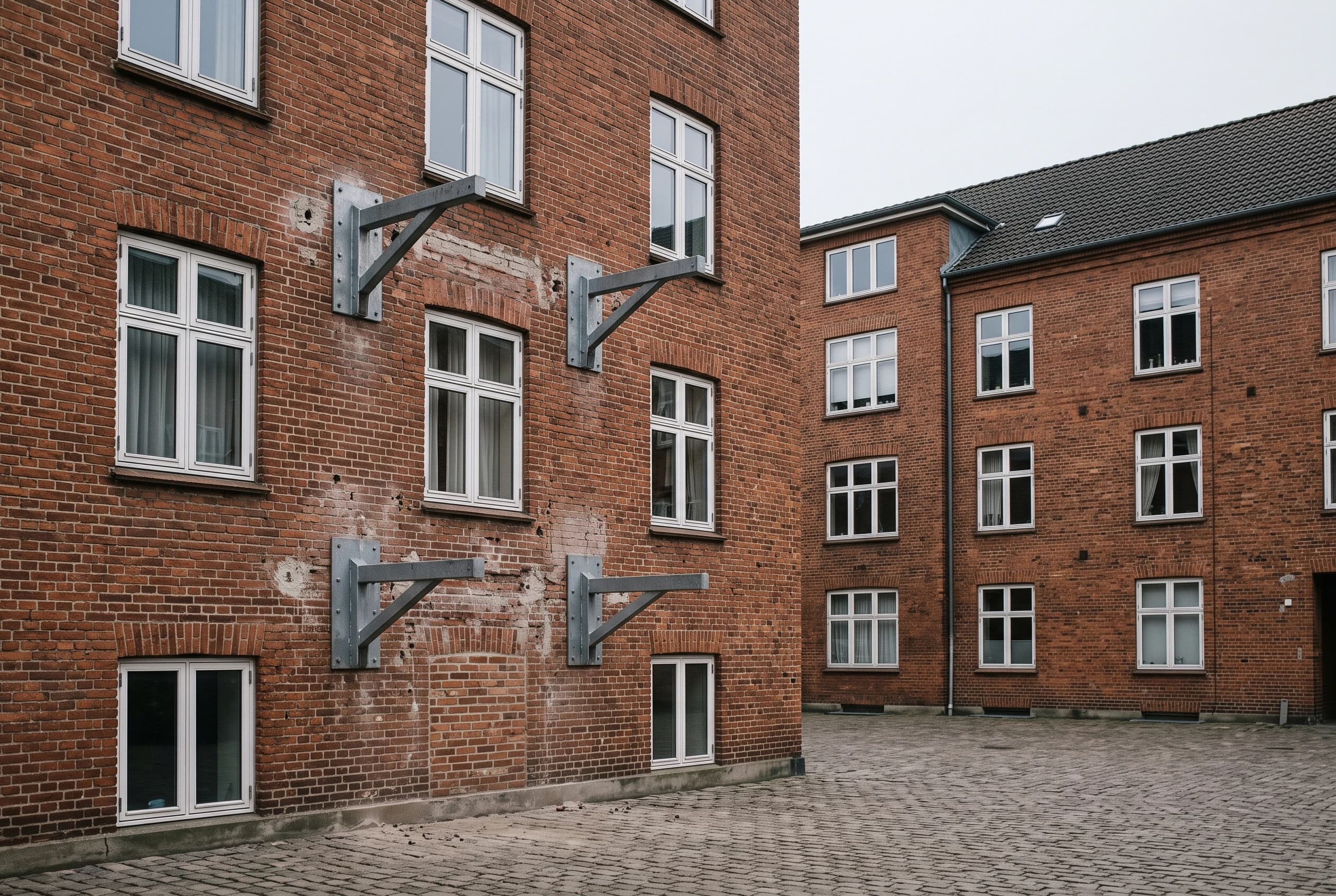 Altankonsoller boltet fast på en rød murstensfacade uden altaner monteret, set fra en brolagt gård en grå dag